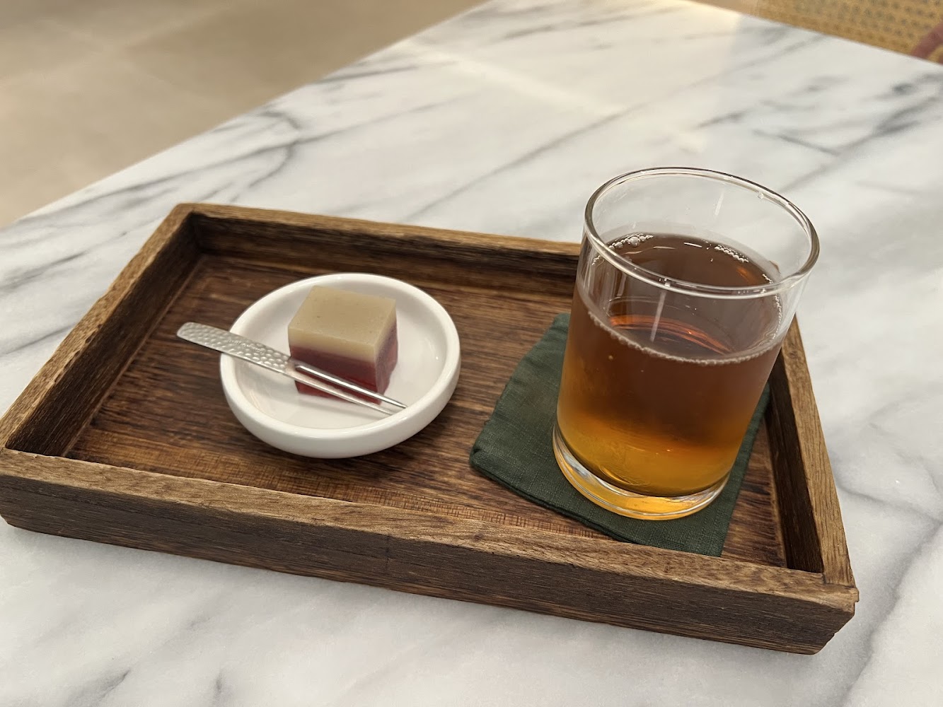這張圖片主要展示一個木托盤，上面擺放著一杯茶飲和一小塊精緻的和菓子，旁邊還有一支小叉子，整體擺設簡約雅致。
