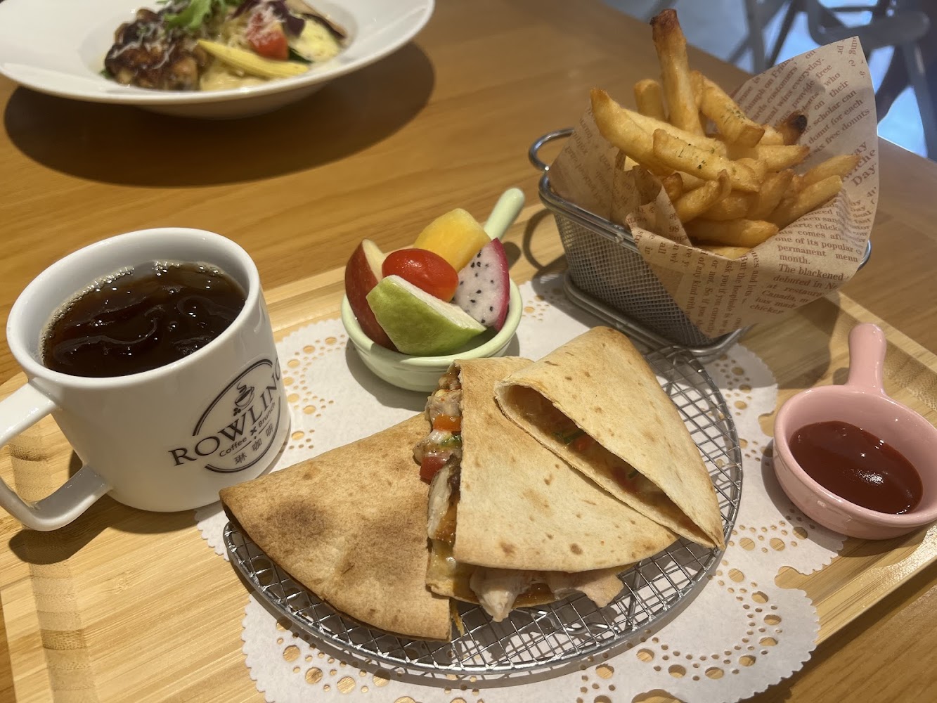 這張圖片主要有一杯黑咖啡、墨西哥餅夾燒烤配料、炸薯條配番茄醬、小碗水果拼盤，食物擺盤精美，是一份豐富的西式餐點。