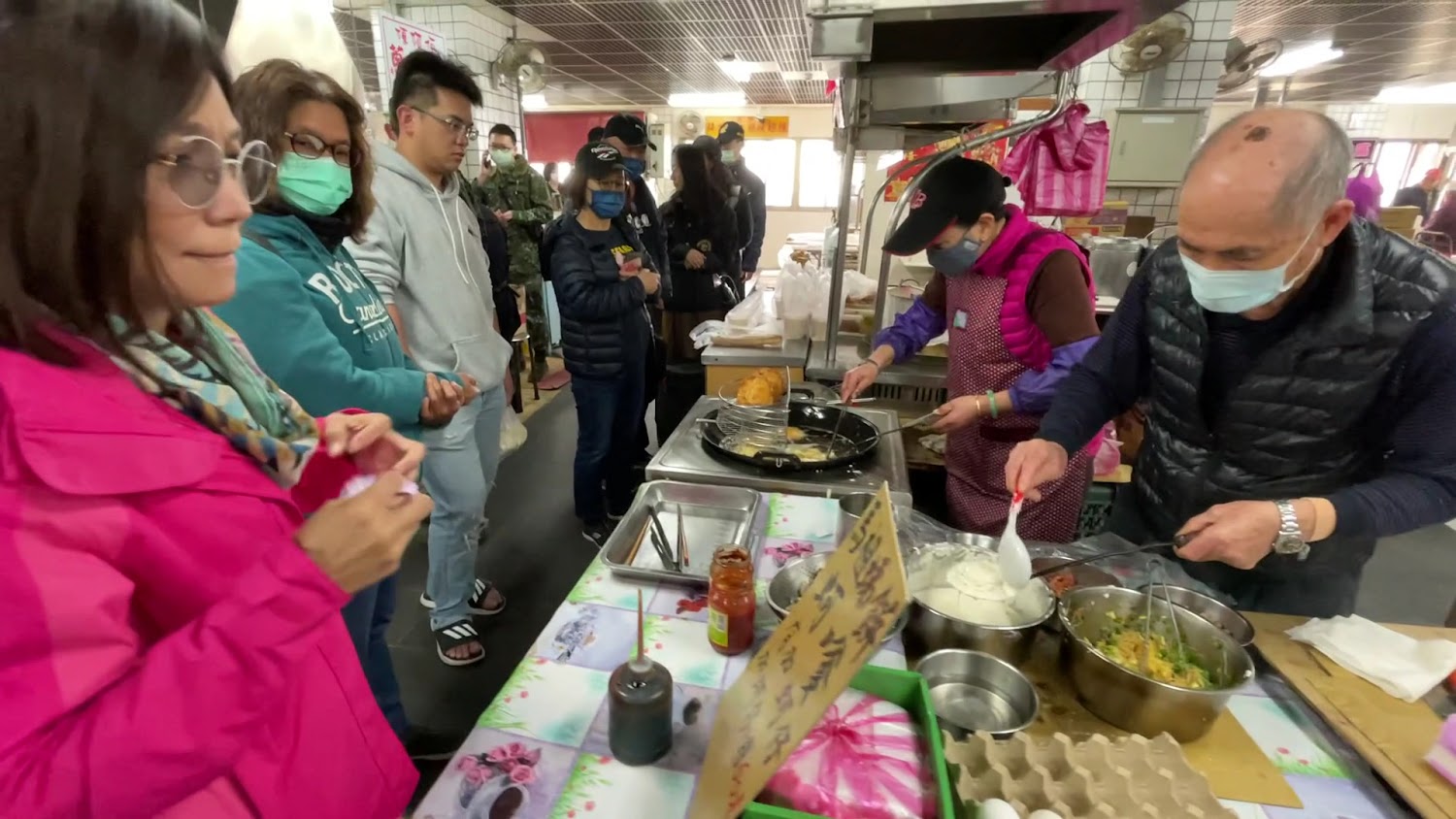 這張圖片拍攝於一間市場的熟食攤，兩位店家正忙著製作料理，前方排有許多戴口罩的顧客等待購買，看起來生意非常興隆，現場氣氛熱鬧。