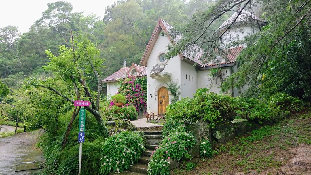 這張圖片主要是一棟童話風格的白色小屋，四周被翠綠樹林和花草包圍，門口有石階和木門，顯得非常幽靜且充滿自然氣息，旁邊還有指示牌。