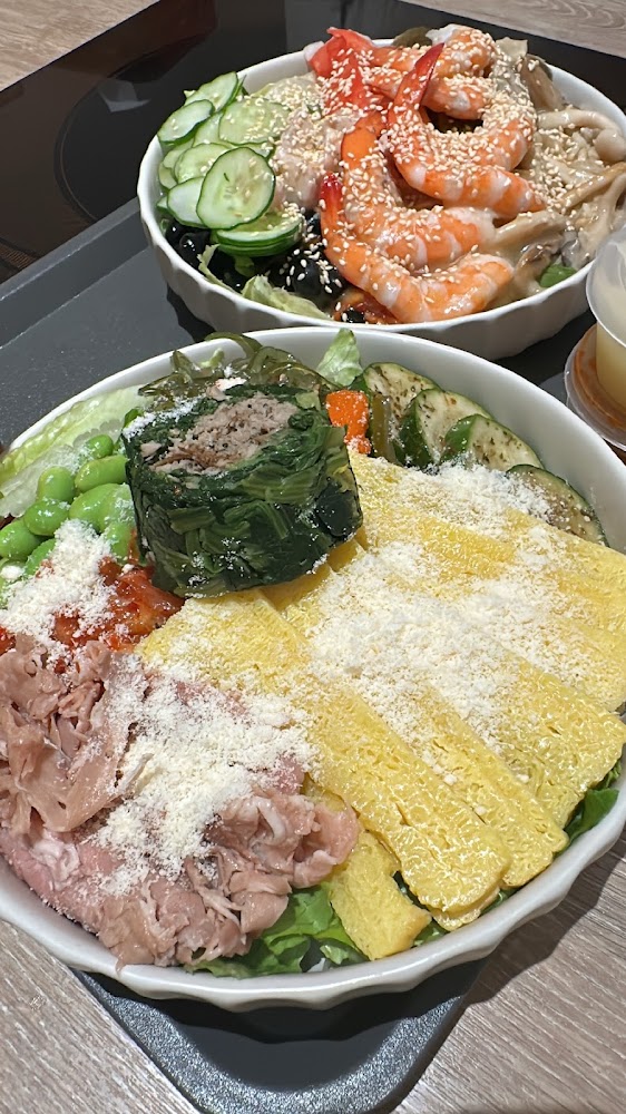 這張圖片展示了兩碗豐富的沙拉丼飯，前方有蛋皮、肉絲、毛豆、昆布捲等配料，後方則有新鮮蝦仁、小黃瓜和芝麻，色彩鮮豔且食材多樣。