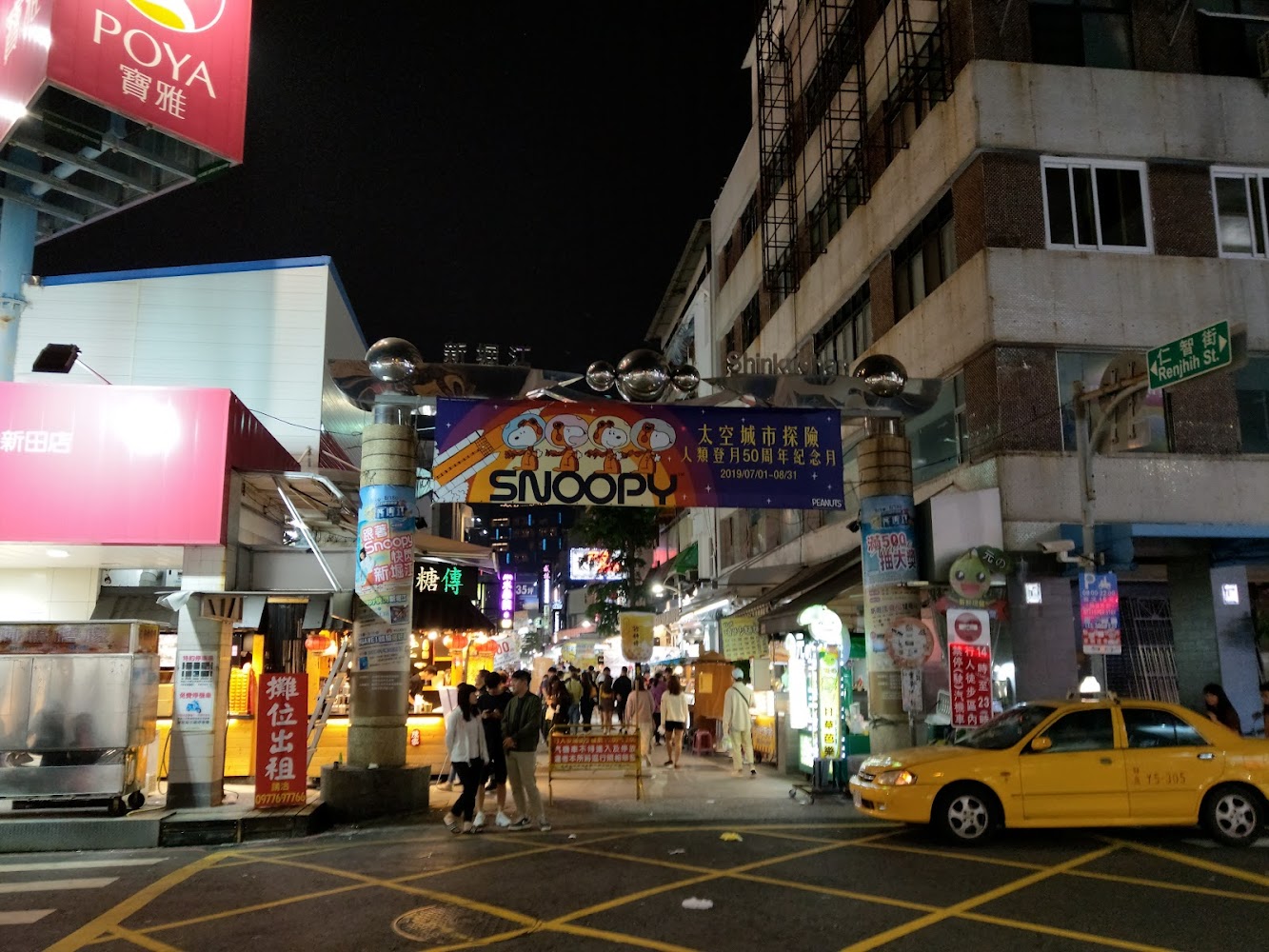 夜晚城市街景中央有熱鬧夜市入口與SNOOPY橫幅，行人來往熱絡。