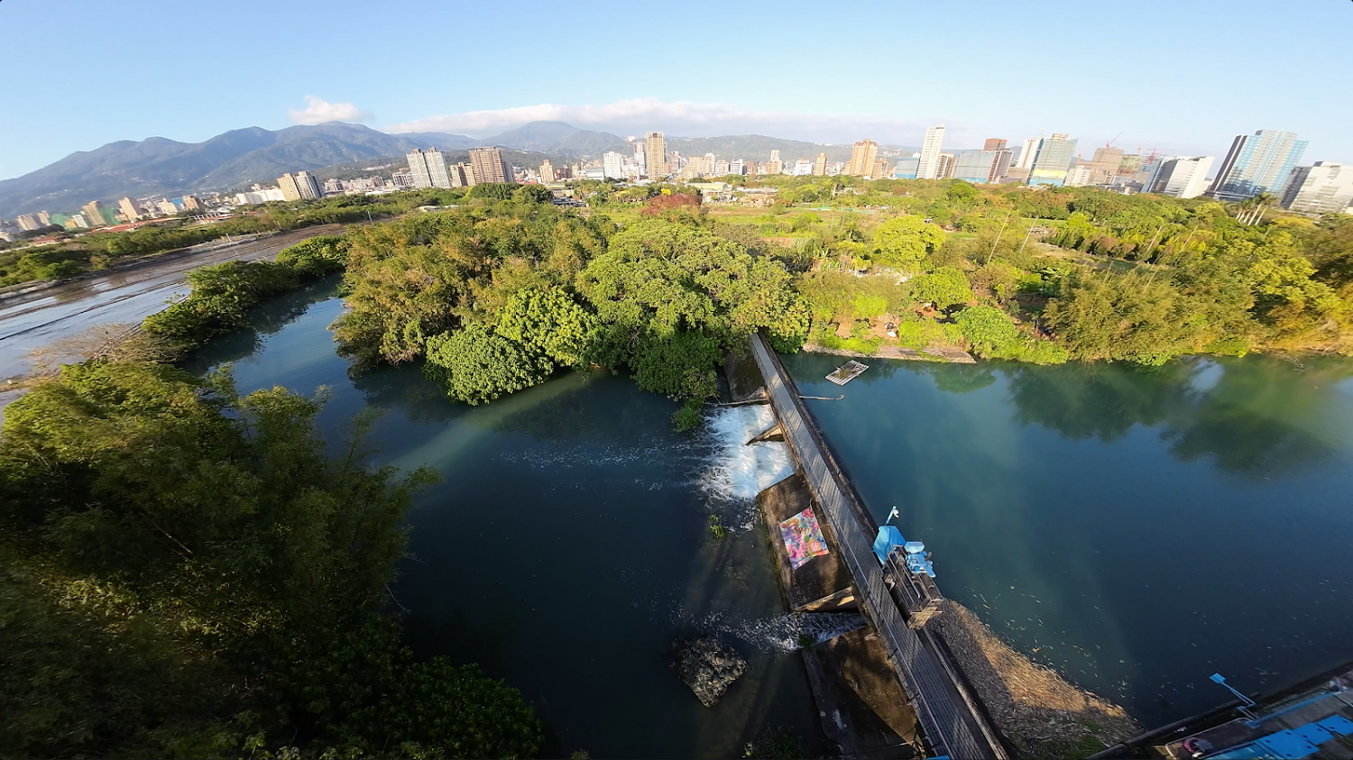 溪流旁綠樹林立遠方有都市高樓與山景中央有小堰壩
