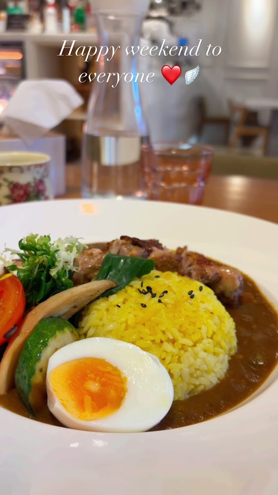 這張圖片展示了一份咖哩飯，配有黃薑飯、半熟蛋、烤雞肉及多種蔬菜，餐點色彩豐富，看起來十分美味。背景為溫馨的餐廳環境，文字祝福大家週末愉快。