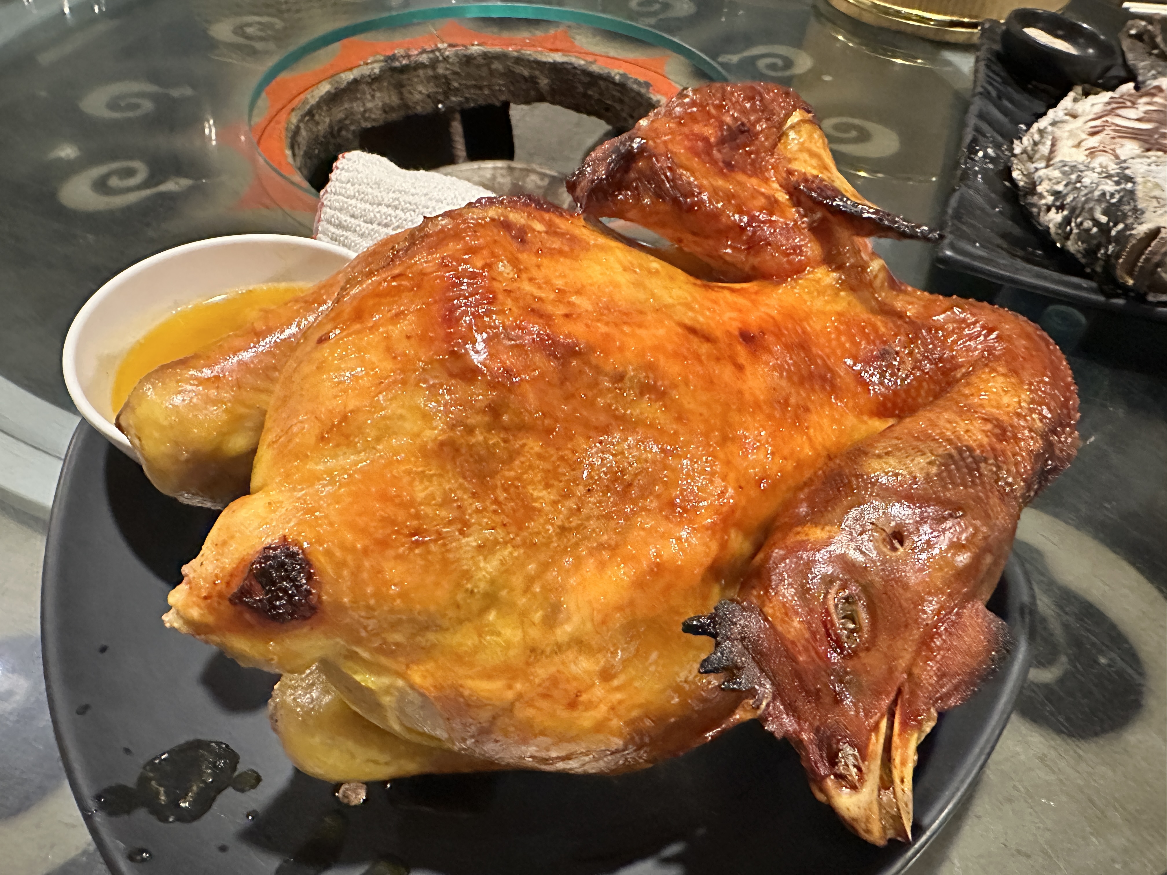 金黃酥脆的烤全雞，旁邊搭配一碗雞汁，擺放在黑色餐盤上