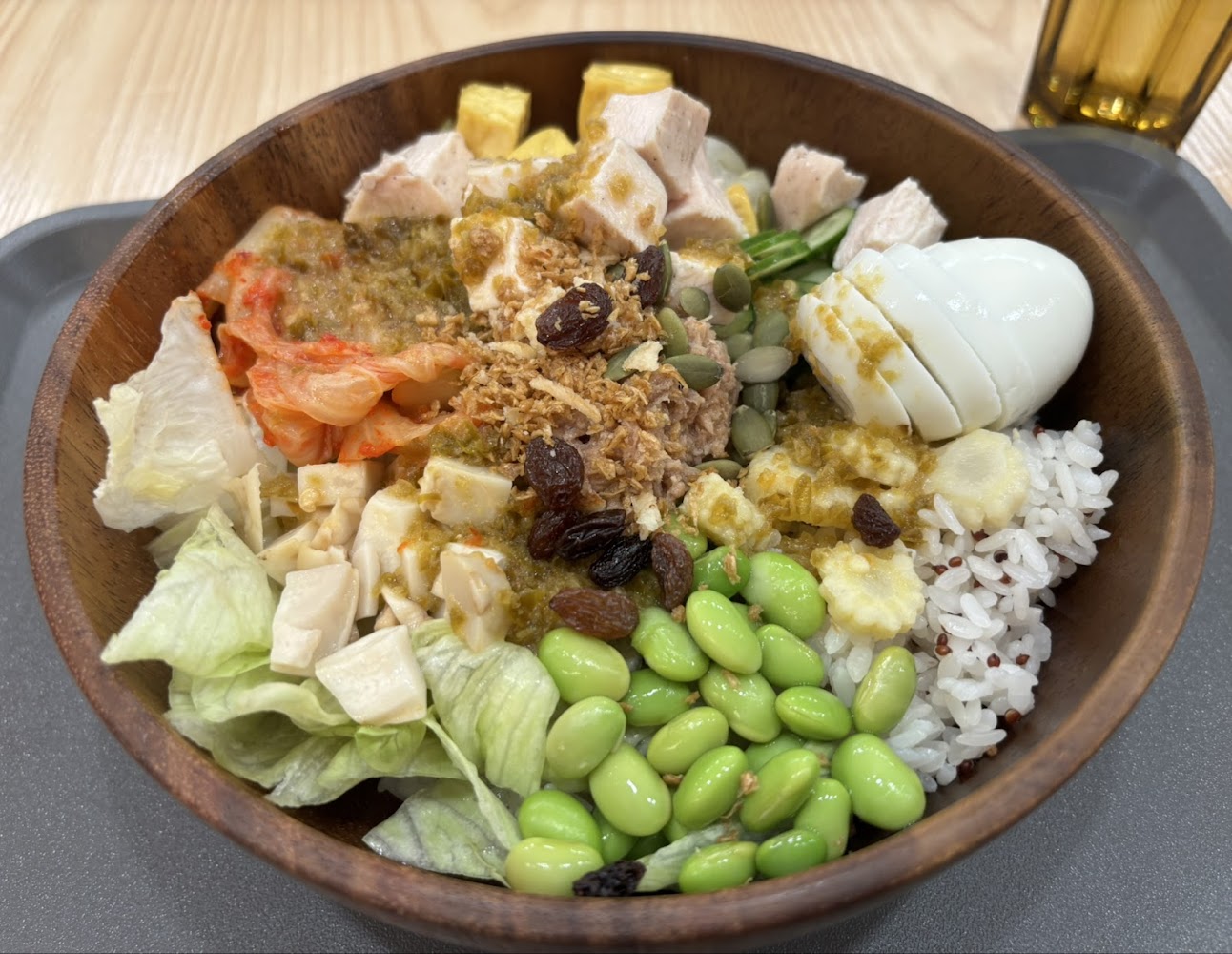 這是一碗豐富的健康拌飯，內含白飯、糙米、毛豆、萵苣、玉米筍、溏心蛋、雞胸肉、泡菜、起司、堅果、葡萄乾以及不同醬料和脆片，搭配一杯飲料，整體營養均衡。