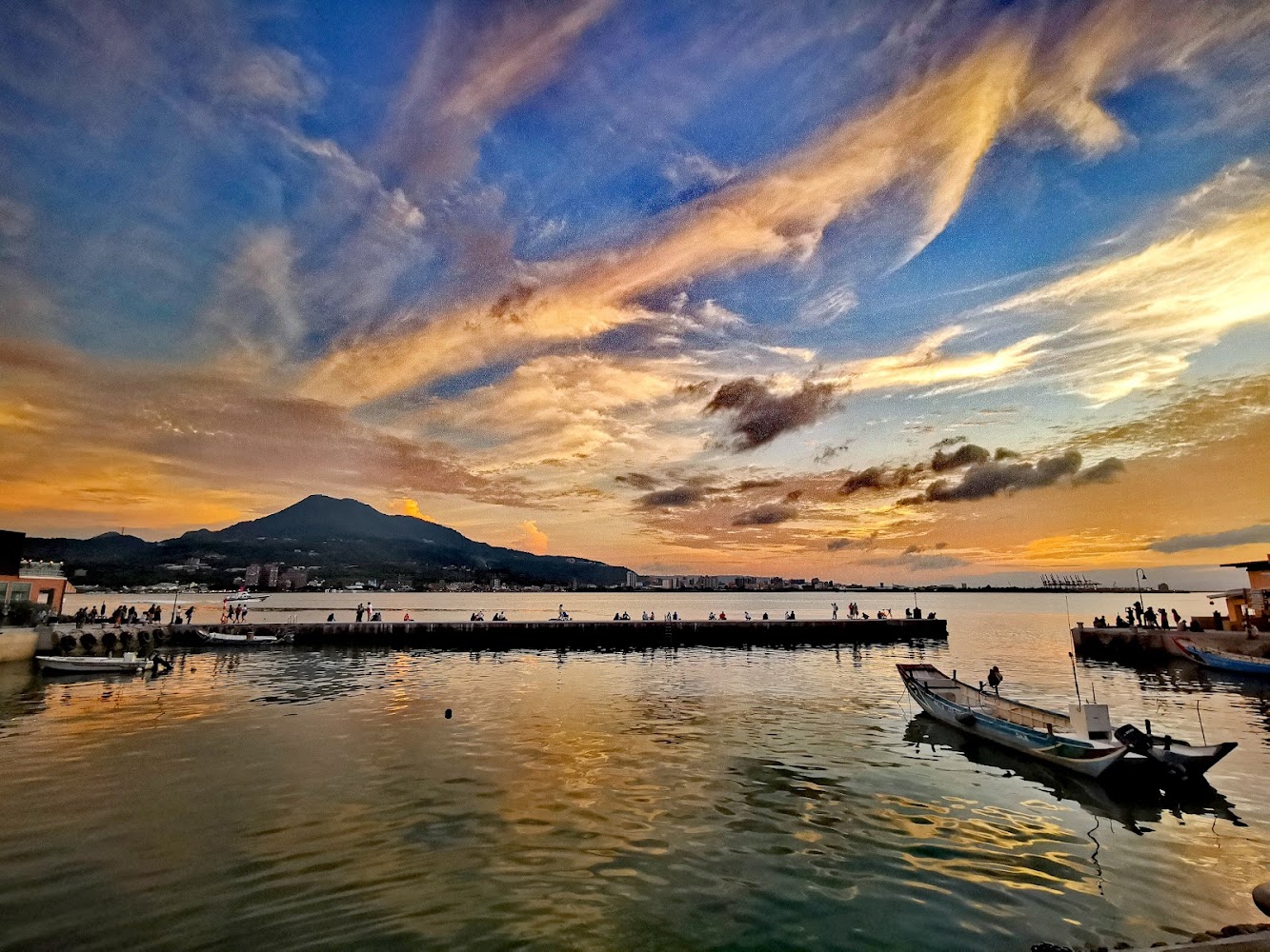 夕陽時分的港口景色，有晚霞、遠山、停靠船隻與釣魚的人們