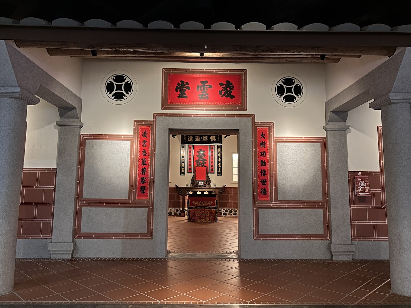 這張圖片展示了一間傳統祖堂，中央有供桌及祖先牌位，四周掛有紅色的對聯與匾額，空間擁有古典門柱與磚牆，氣氛莊嚴典雅，承襲中華文化的傳統氛圍。