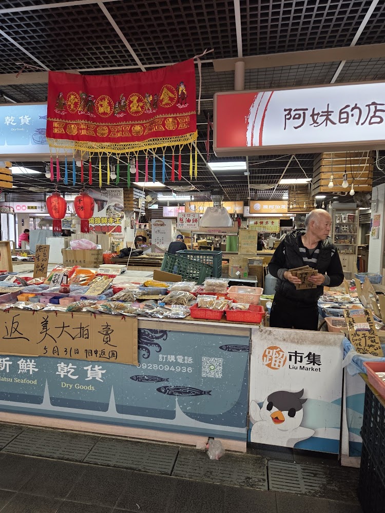 這張圖片拍攝於傳統市場，畫面中有販賣各類乾貨與海鮮的攤位，桌上擺滿包裝食品。上方掛有傳統紅色布條裝飾，旁邊有燈籠與明顯的市場招牌，展現熱鬧的市場氛圍。