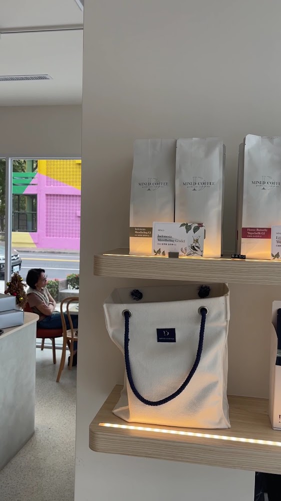 這張圖片主要展示了一間咖啡店的商品展示架,上面有MIN.D COFFEE品牌的咖啡豆包裝和一個白色帆布袋,背景能看到玻璃窗外鮮豔彩色的建築和店內休息的客人。