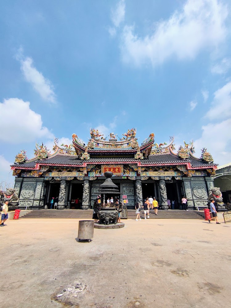 一座傳統華人寺廟，建築裝飾精美，屋頂有雕刻和彩繪，前方有大香爐，許多人進出寺廟，場地寬敞，天氣晴朗。