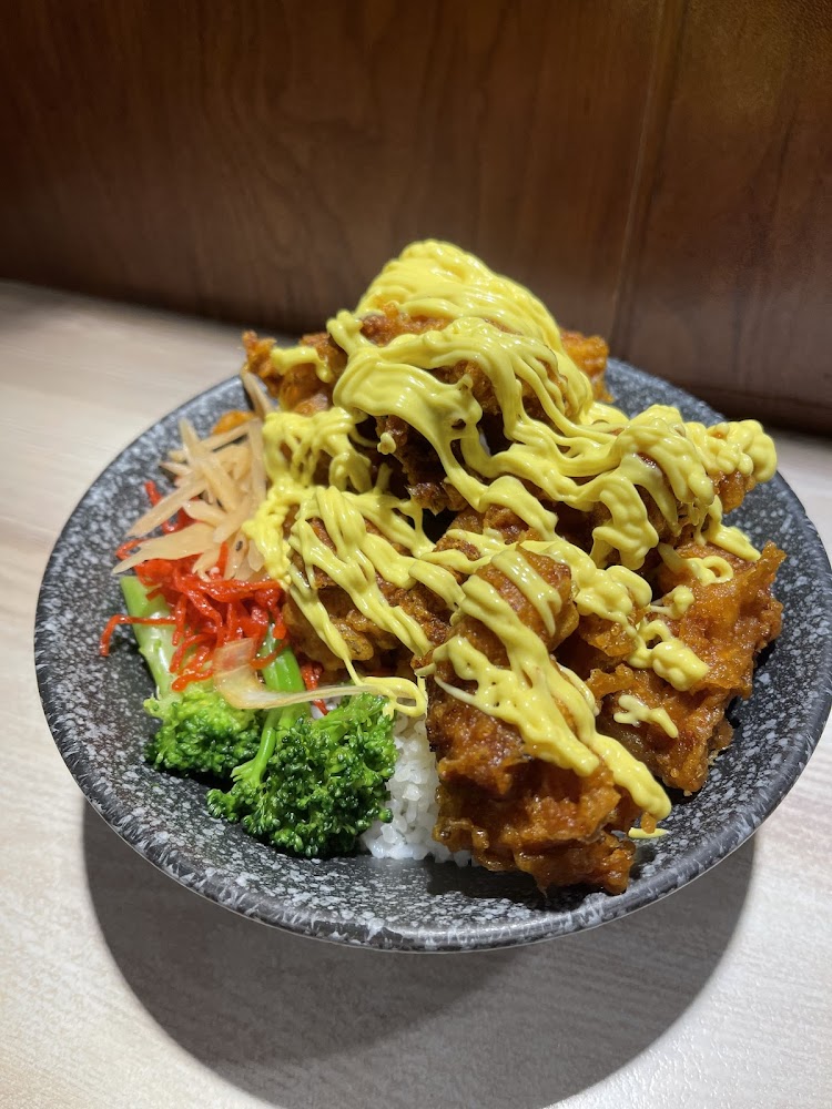 這張圖片裡有一碗白飯，上面放著炸雞塊並淋上黃色醬料，旁邊有花椰菜、紅蘿蔔絲和醃製配菜，配色鮮豔，看起來十分美味。