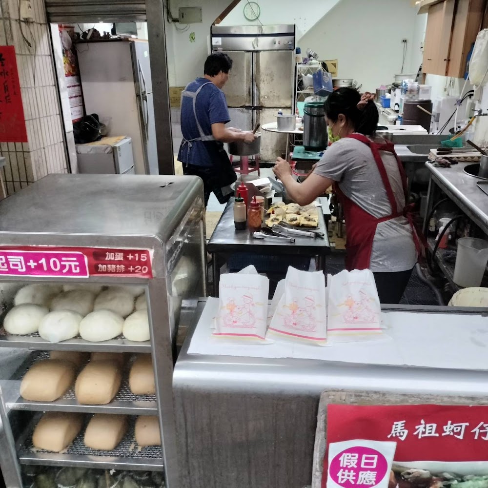 這是一間傳統早餐店，前方有蒸包、饅頭等食品，桌上有包裝袋，店員正在準備餐點，環境樸實，呈現忙碌的工作氣氛。