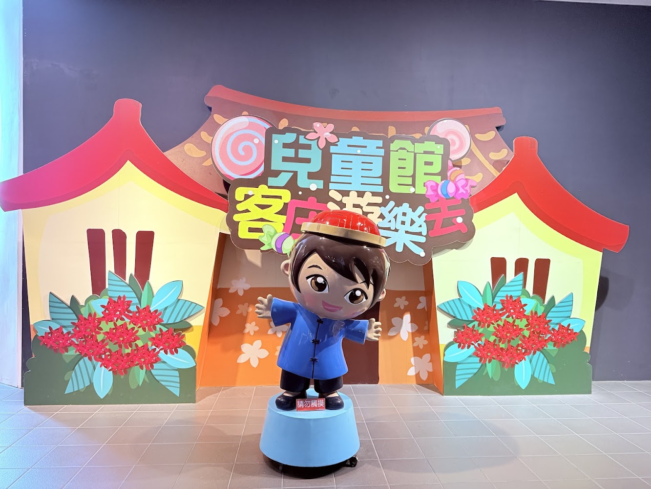 這張圖片展示了一個卡通人物雕像，穿著藍色衣服和紅色帽子，前方寫有『請勿攀爬』。背景是以客家建築和花朵為主題的裝飾板，上方有『兒童館 客家遊樂去』的彩色字樣。