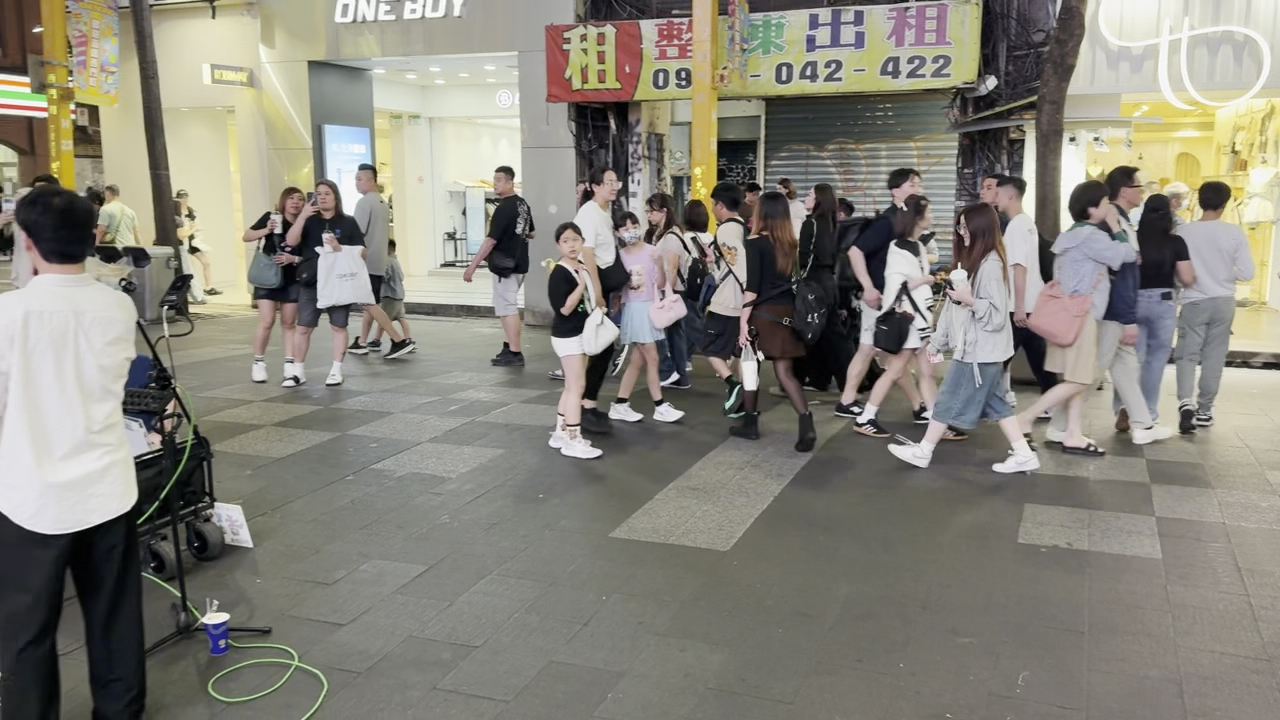 西門町徒步區夜晚街景，街頭表演者左方演唱，右側人群熙攘，兩旁時尚服飾商店燈火通明