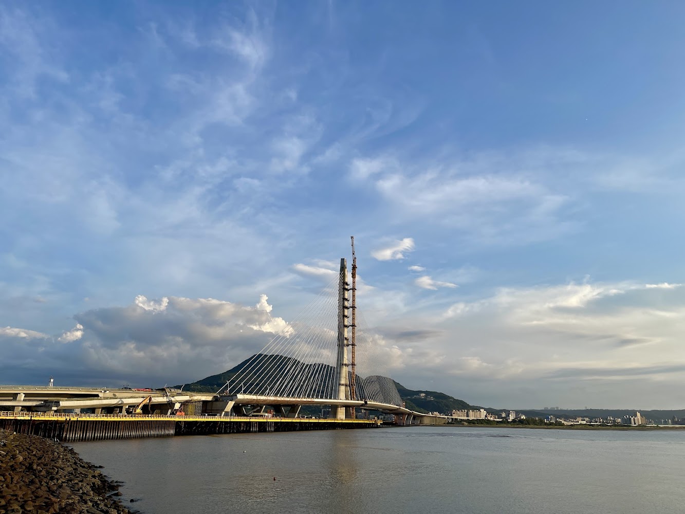 這張圖片以夕陽下的景色為主，前景是一座正在建造中的斜張橋，橫跨在河流之上，背景可見青山與城市建築，天空蔚藍且有白色雲朵。