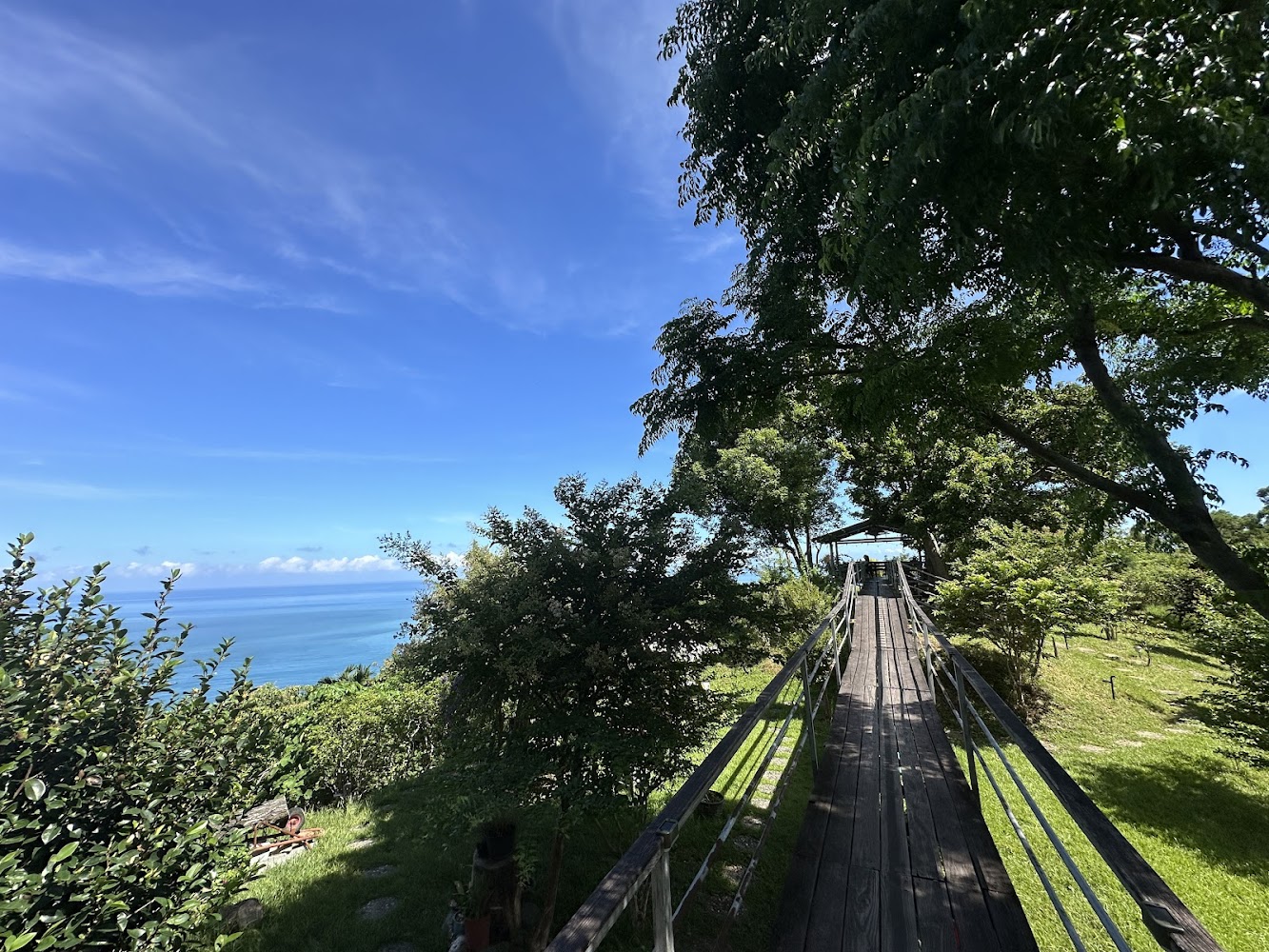這張圖片展示了一條木棧橋，兩側被綠樹環繞，遠方可以看到蔚藍的海洋和晴朗的天空，整體景色自然清新，充滿悠閒氣氛。
