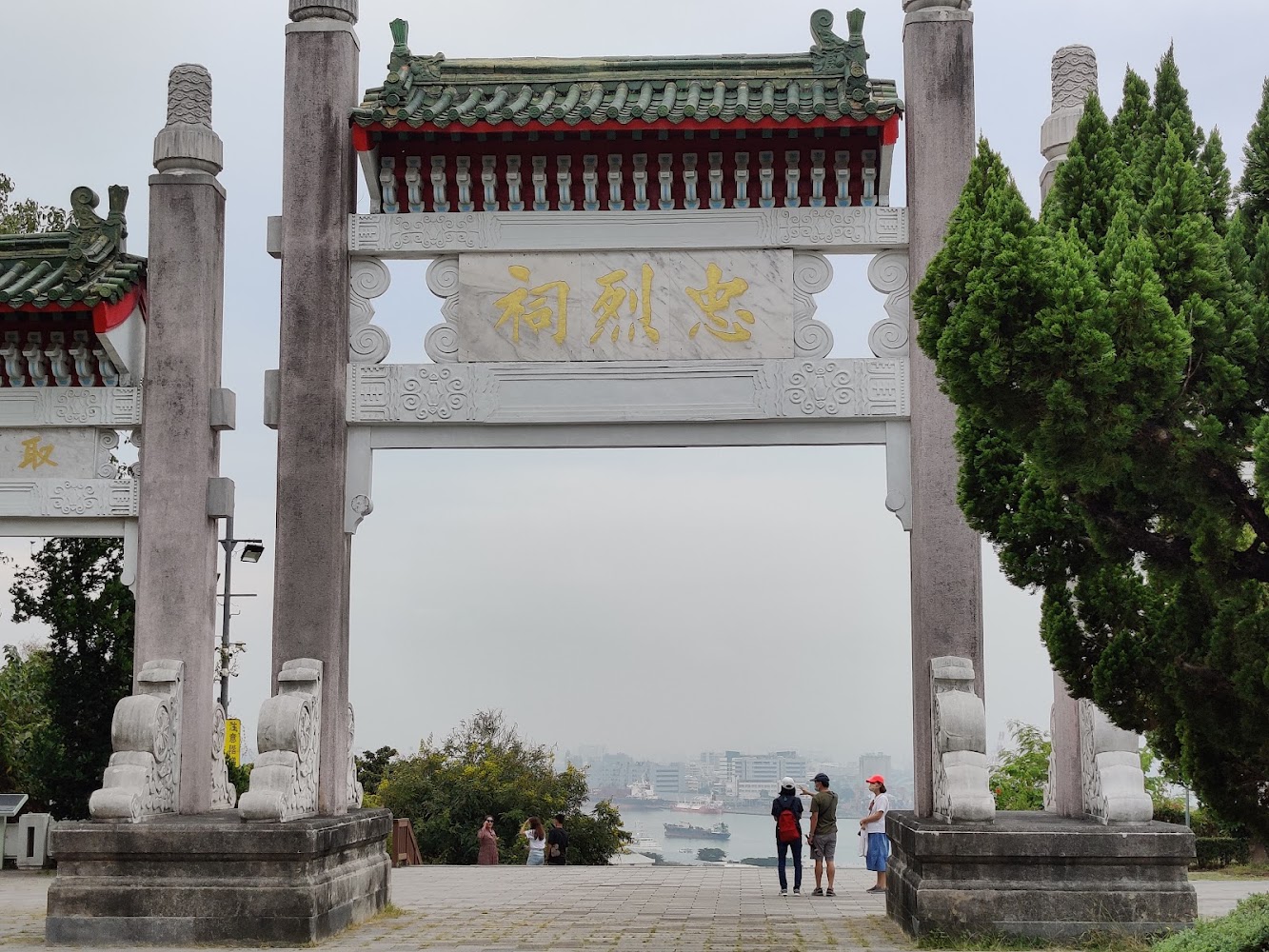 傳統中式牌樓寫有忠烈祠，背景可見城市與海港景色。