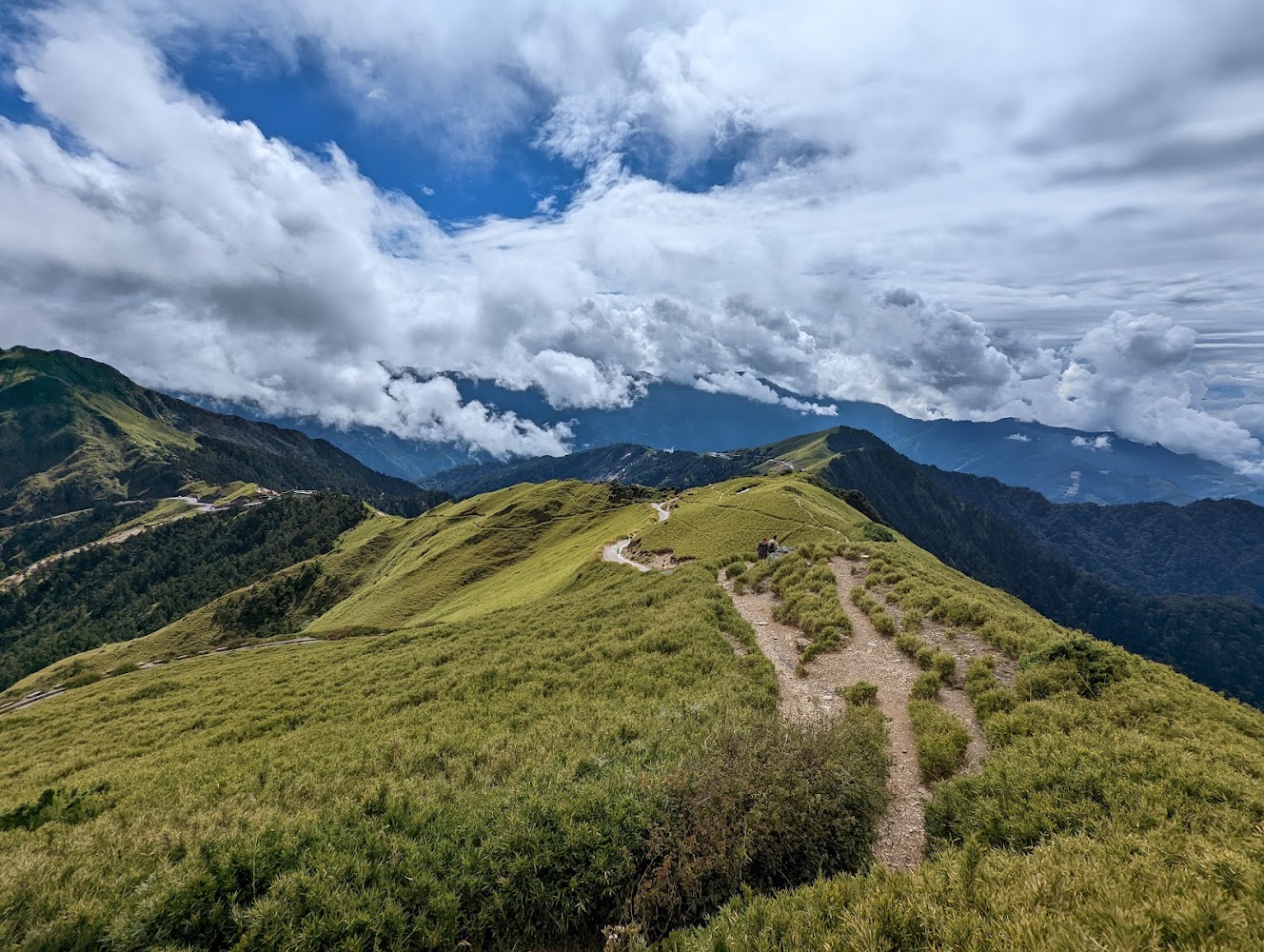 這張圖片主要呈現壯闊的高山景色,綠色草原與蜿蜒的登山步道延伸至遠方,天空滿布雲朵,層次分明,整體畫面十分遼闊壯麗。圖片無明顯浮水印。