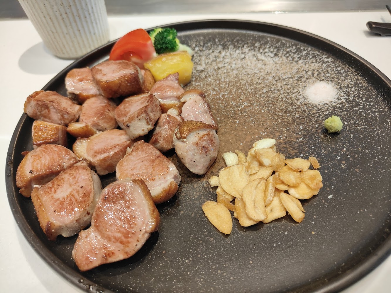這張圖片主要有切塊的烤鴨肉、蒜片、鹽、哇沙比，以及配菜如番茄、花椰菜和馬鈴薯，擺放在黑色餐盤上，看起來精緻可口。