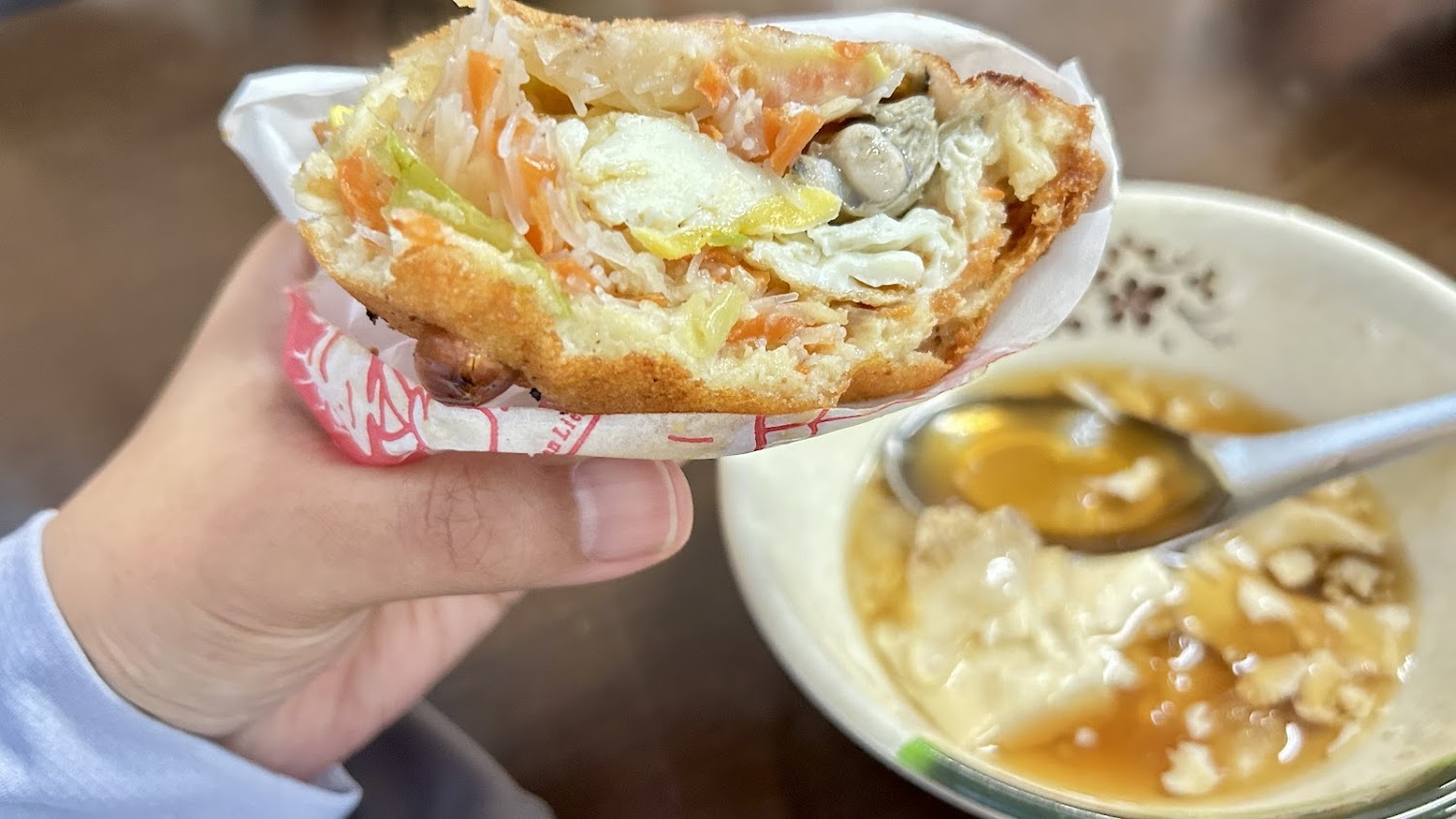 手拿炸餅斷面可見蔬菜與蚵仔，旁邊有蛋花湯