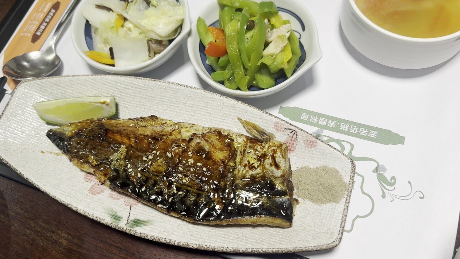 這張圖片主要有一份日式烤魚排，搭配檸檬片和胡椒鹽，旁邊有涼拌青椒、燙蔬菜和一碗清湯，是一套餐式健康餐點。
