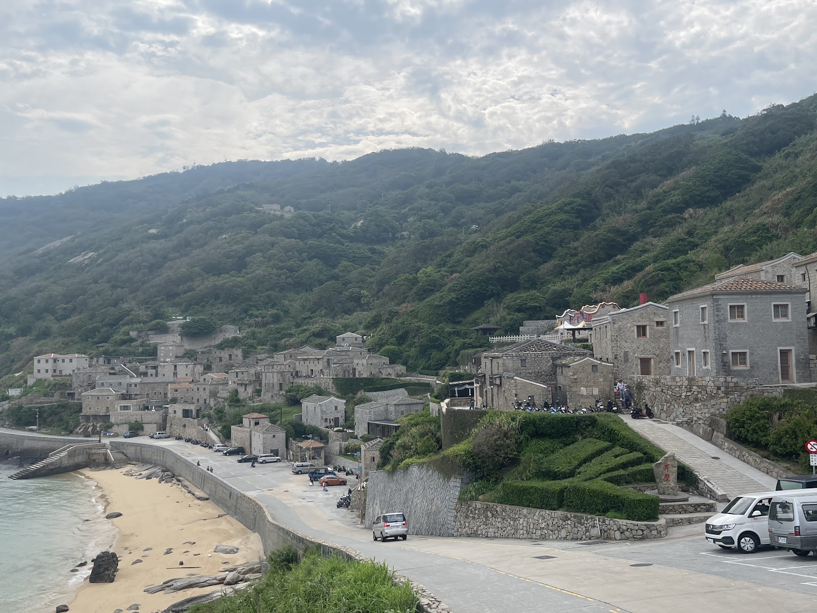 芹壁聚落俯瞰，石砌建築群沿山坡延伸，左側弧形海灣沙灘，翠綠山頭為背景