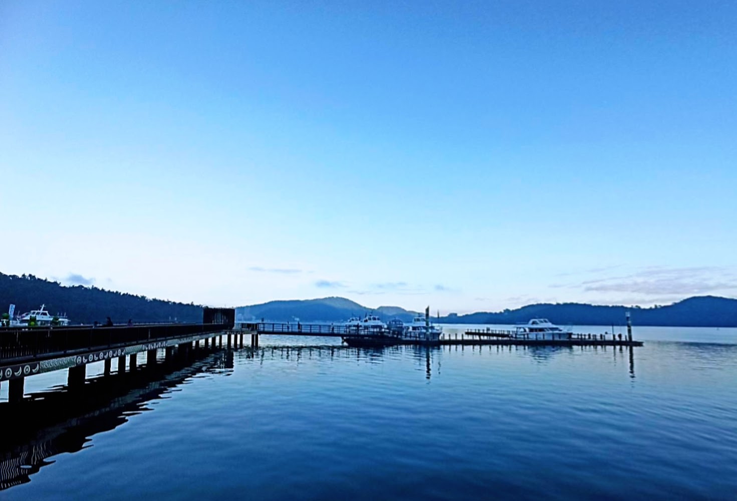 一座碼頭，水面上停靠著幾艘遊艇，背景遠處是青山和寬闊的天空