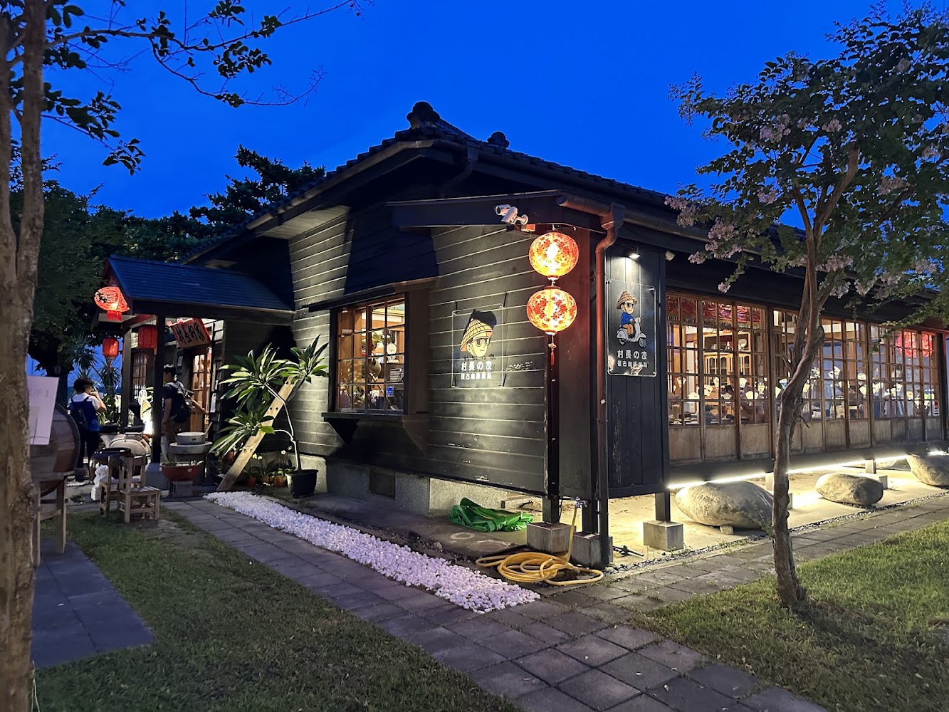 日式風格木造建築，掛著紅色燈籠，室內燈光溫暖且有顧客用餐