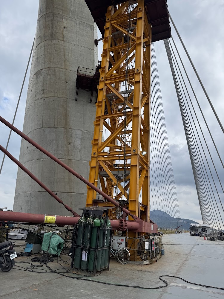 橋樑施工現場有巨大混凝土橋塔、黃色鋼結構施工設備,以及多條鋼索