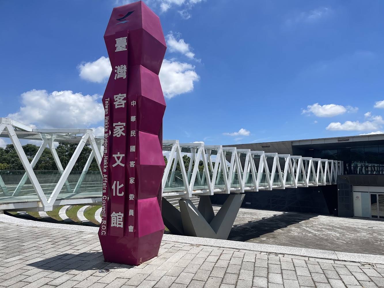 照片中有一座現代設計的建築，旁邊矗立著顯眼的桃紅色指標，上面寫著『台灣客家文化館』，是推廣客家文化的重要場所。