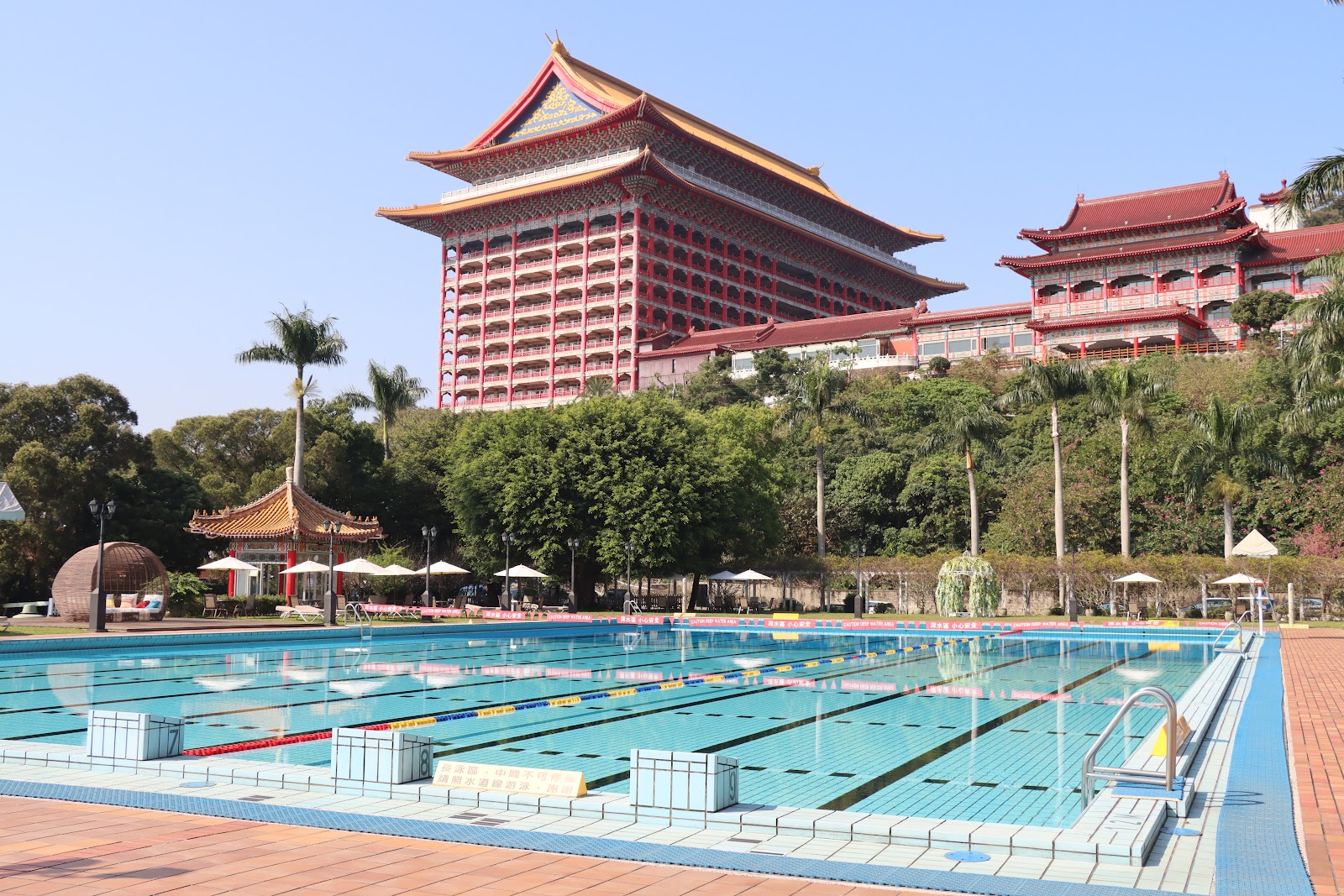 圓山大飯店戶外泳池，清澈藍色水道，背景是宮殿主建築與棕櫚樹