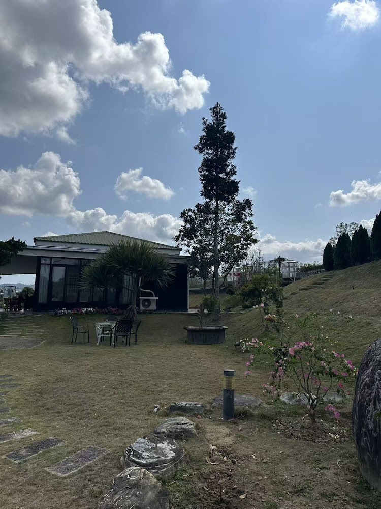 這張圖片主要呈現一個庭院場景，有平坦的草地、零星的花朵、樹木，一旁有座現代建築，還有戶外桌椅。天空湛藍，雲朵飄浮，整體氣氛悠閒自在。圖片中無浮水印。