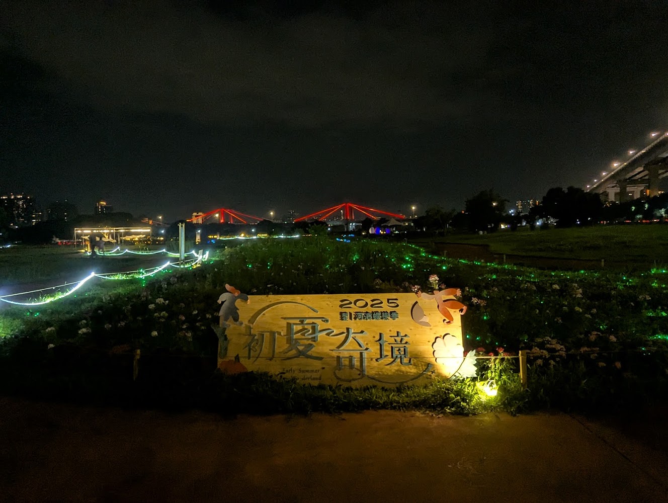 這張圖片是一個夜間的戶外活動現場，有亮燈裝飾的草地和遠處點綴紅光的大橋，前景有寫有『2025 初夏奇境』的大型牌子。