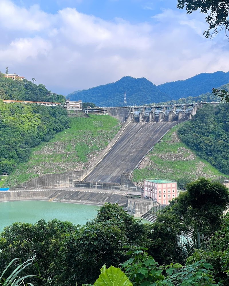 一座大型水壩，水壩下方有一座粉紅色建築，周圍環繞著青山與豐富的綠色植被，前方有碧綠色的水體。