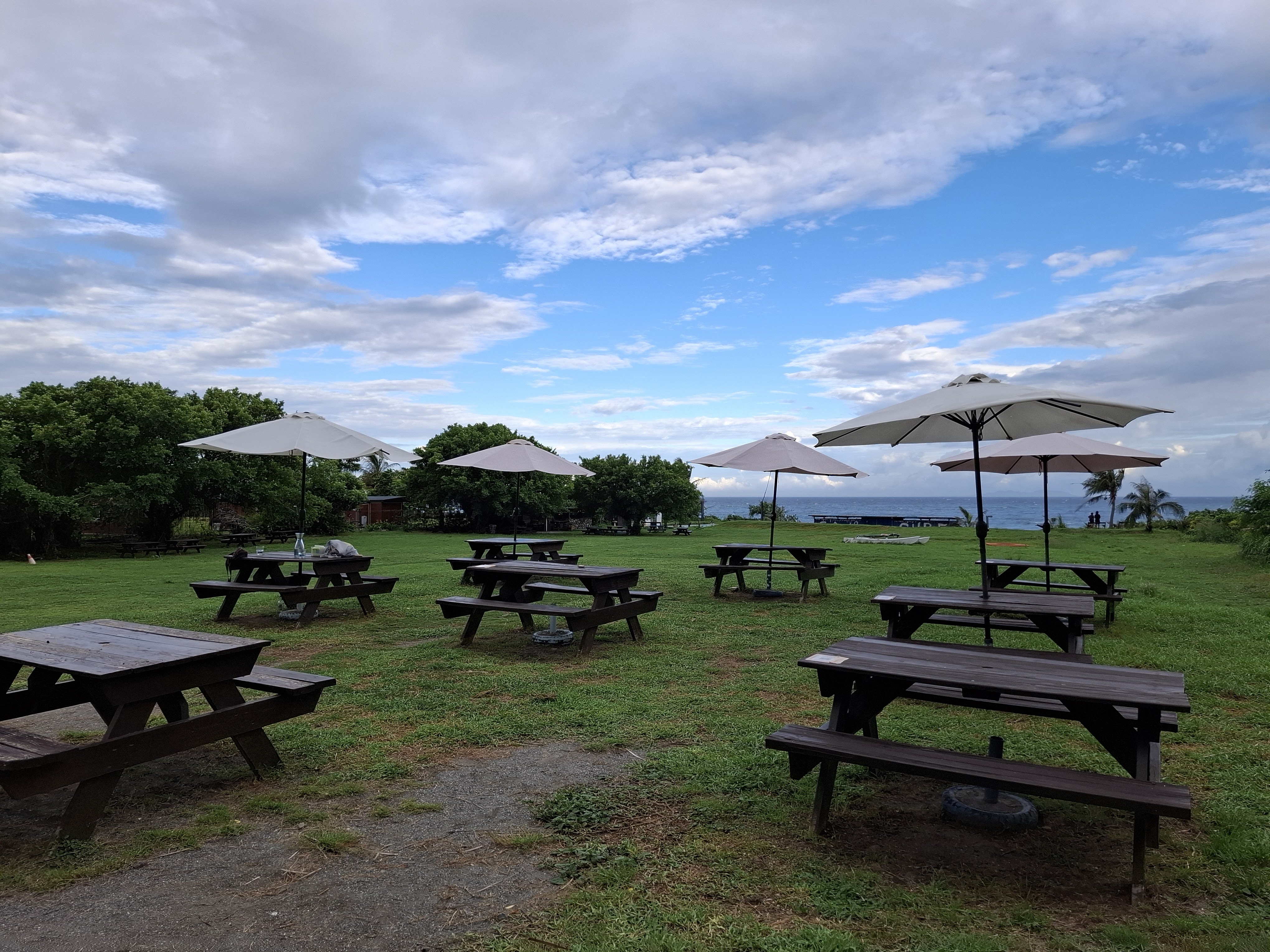 草地上的木製長椅餐桌和白色遮陽傘，遠方可見海景