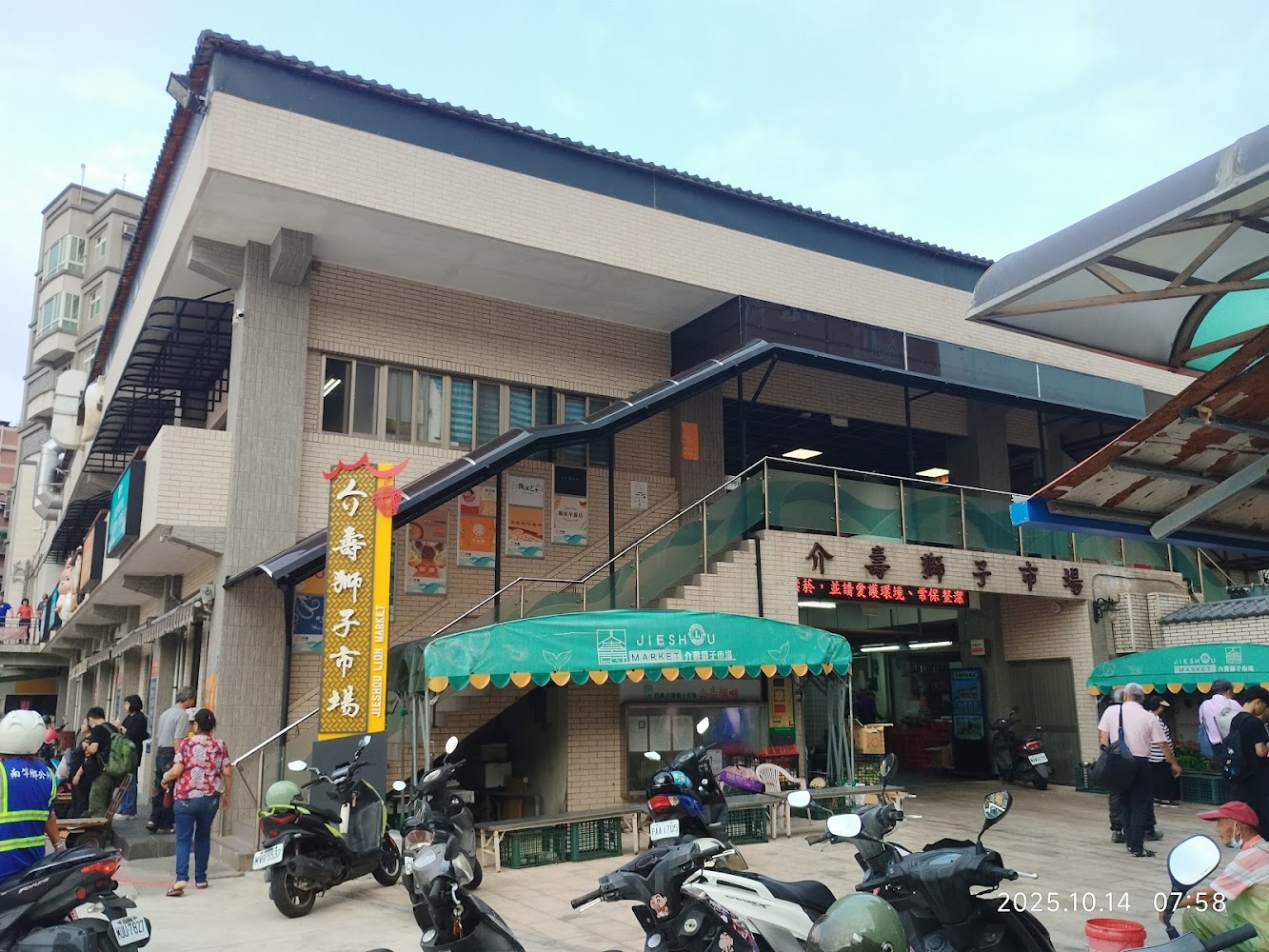 這是一張傳統市場的照片，圖中可見市場建築外觀、人們進出、市場入口招牌及停放的機車，展現熱鬧的生活氛圍。