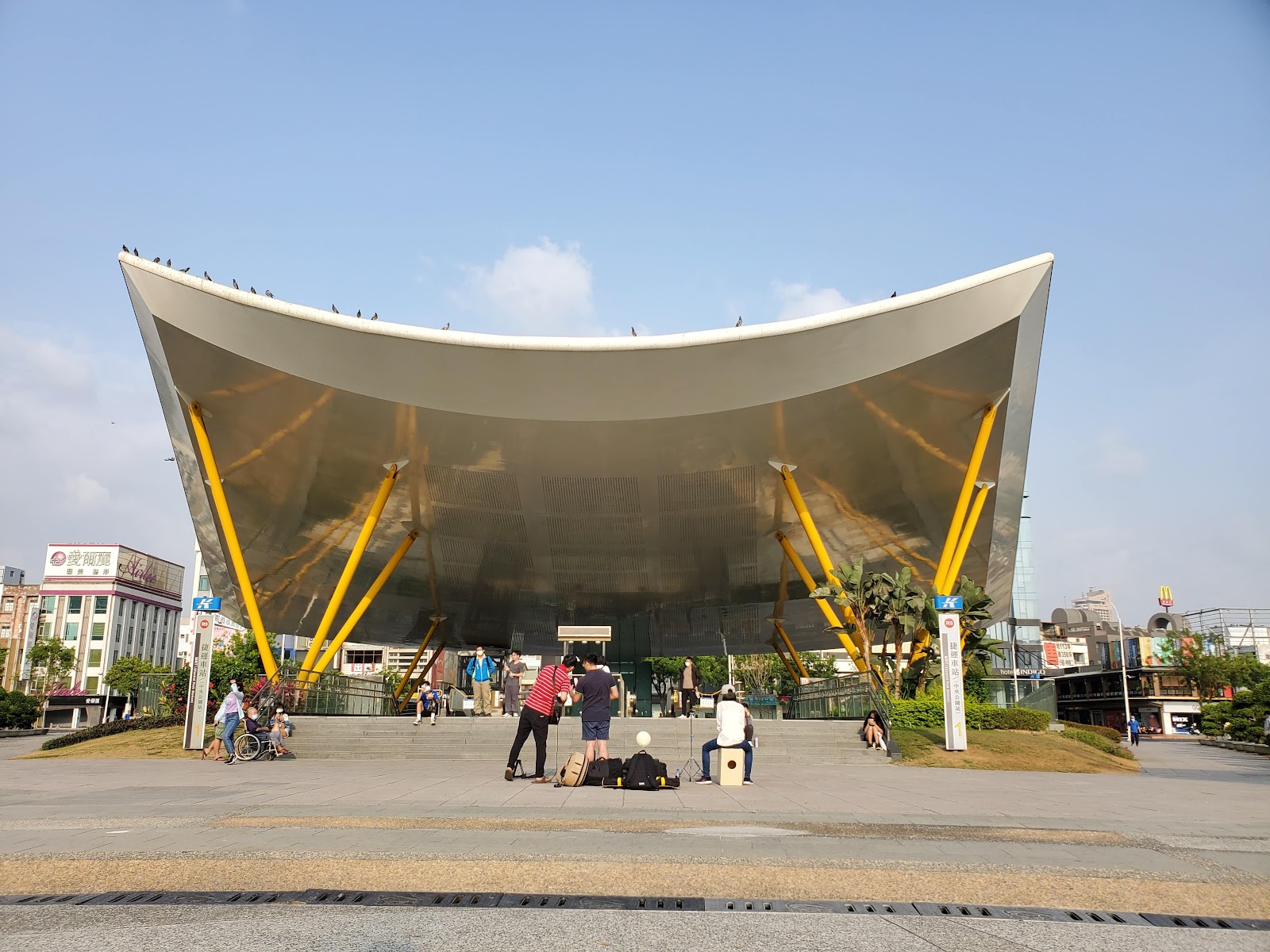 高雄中央公園站地面出口，白色弧形遮棚與黃色柱體在藍天下格外搶眼