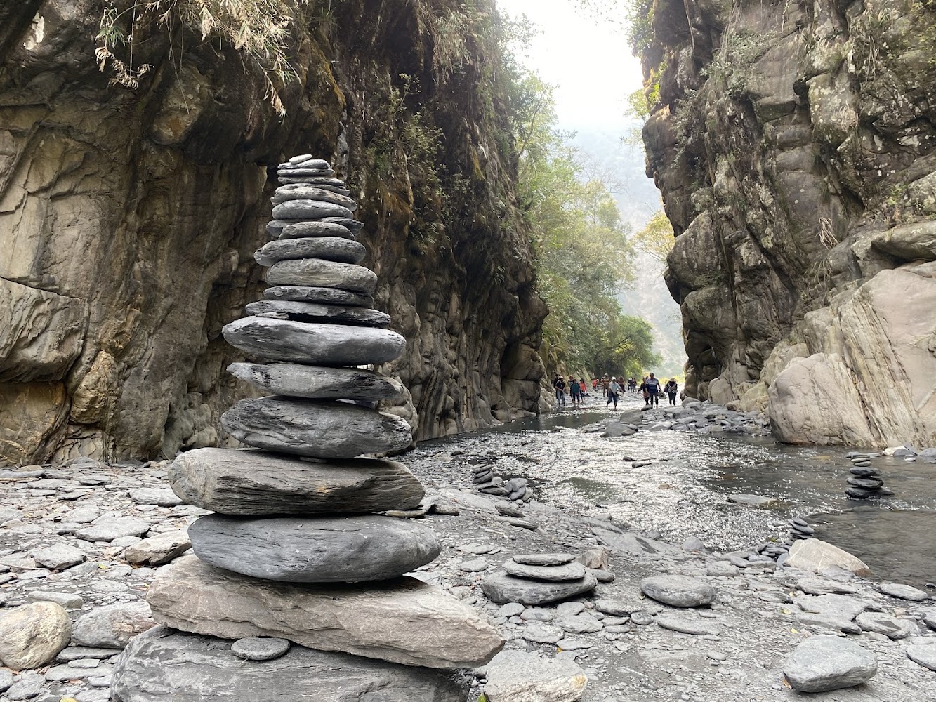 溪流旁有平衡石堆疊，後方有峽谷與穿越溪流的遊客