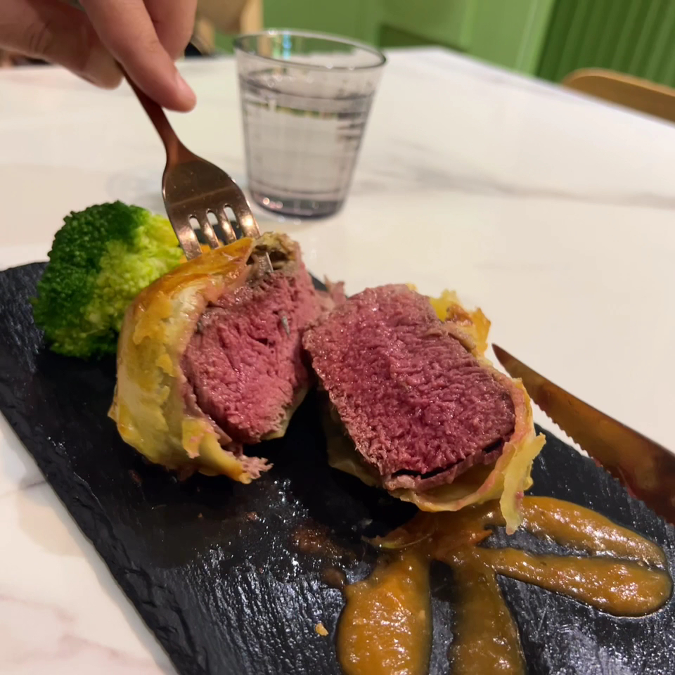 圖片中是一道切開的威靈頓牛排，牛排外層包裹著酥皮，內部肉質粉嫩，旁邊有花椰菜和醬汁，桌上還有一杯水。