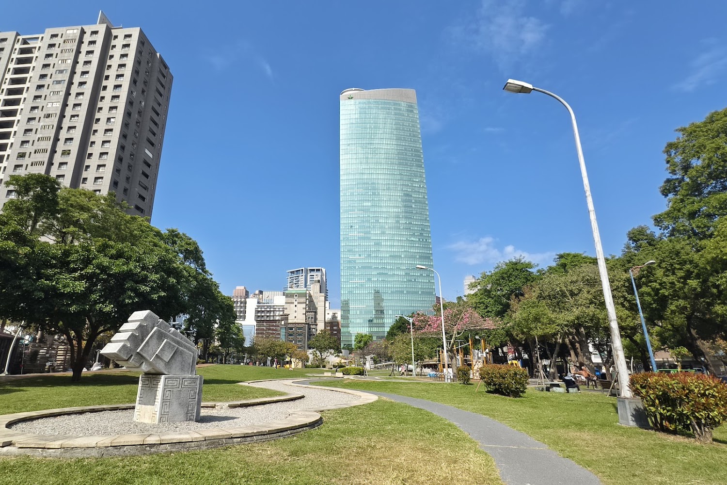 高聳玻璃大樓前方有公園綠地、雕塑與行人步道