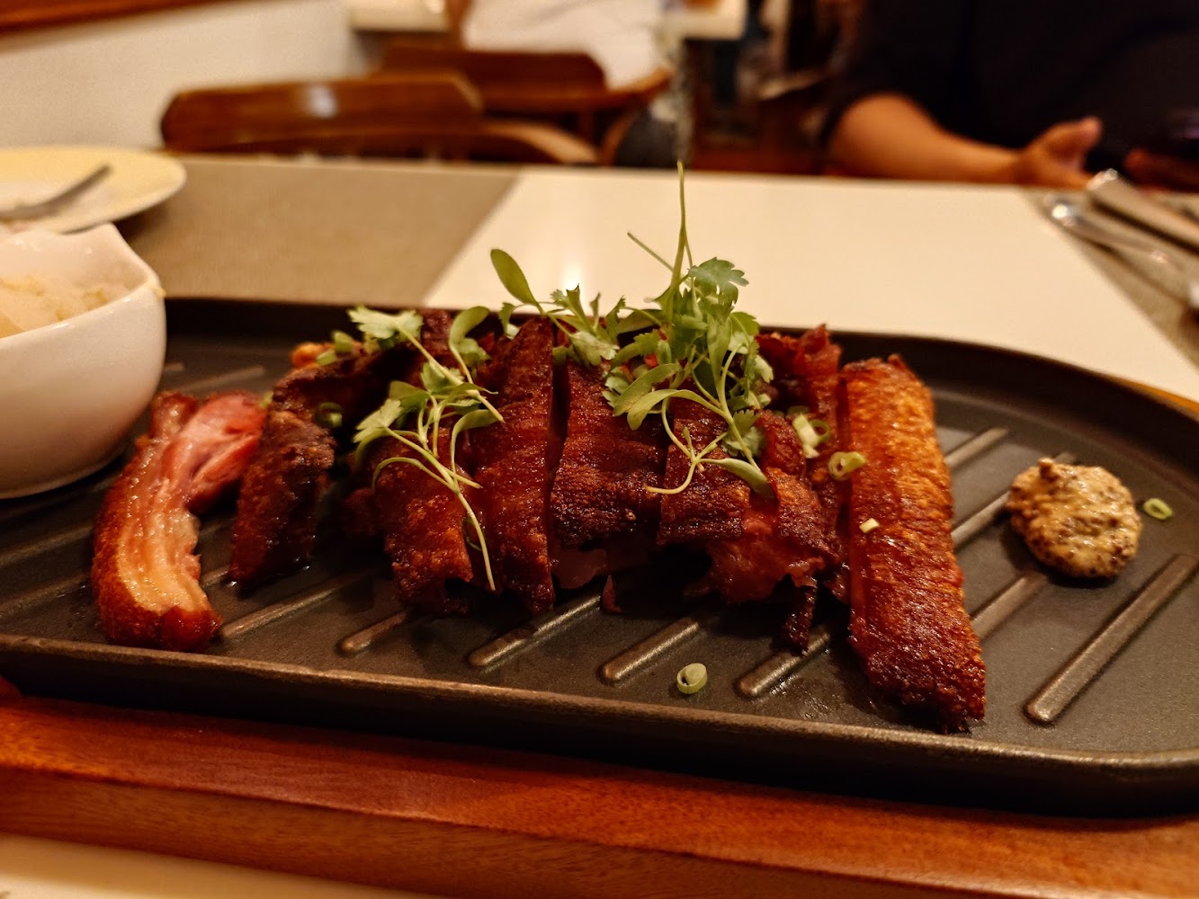 一道烤肉主菜，肉塊外皮酥脆，上面撒有新鮮香草和蔥花，旁邊附有芥末醬和一碗配菜