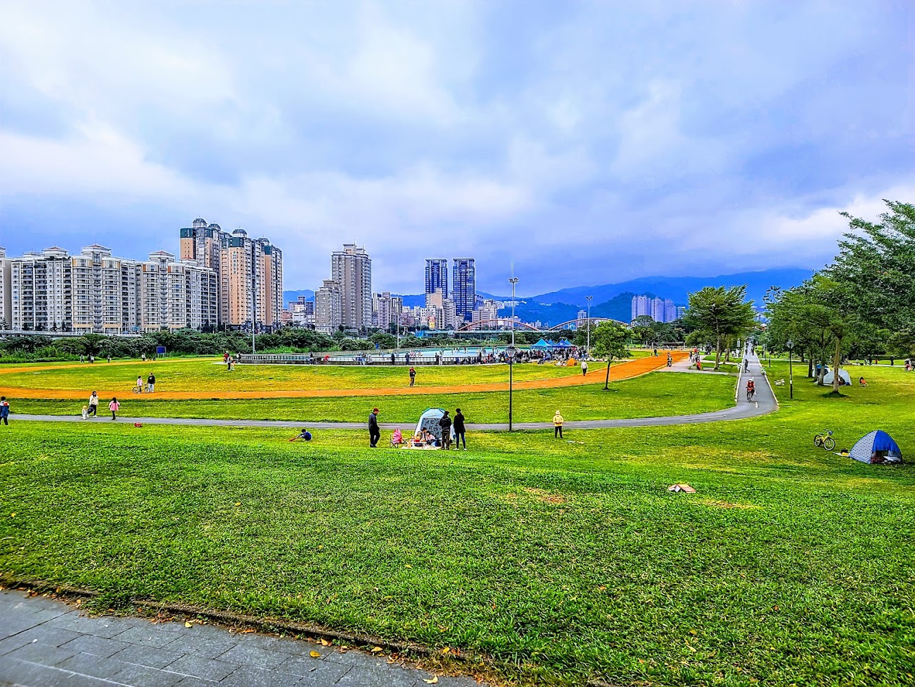 城市公園大片綠地與步道，遠處可見高樓與山景
