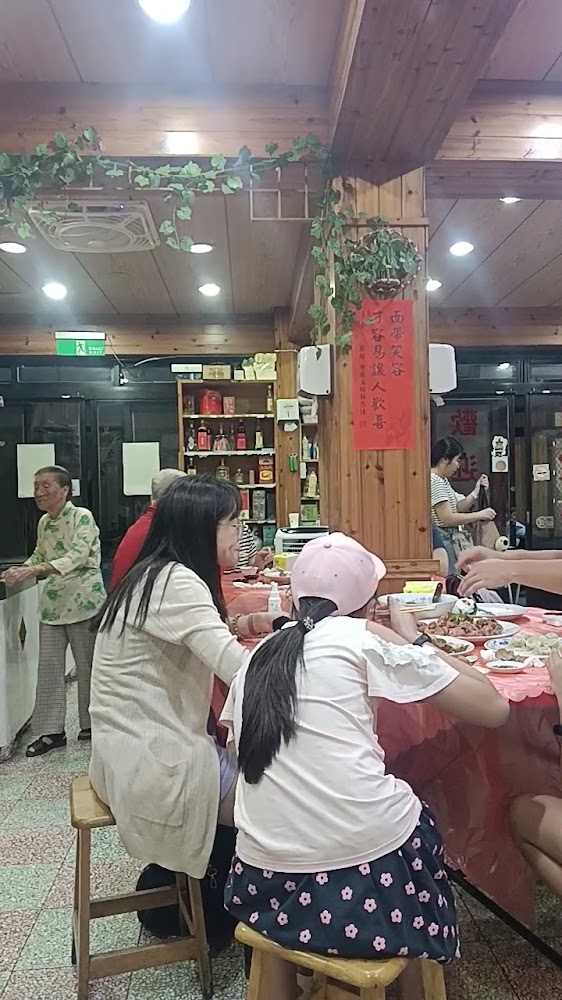 這張圖片主要是在一間中式餐廳，大家坐在圓桌前享用美食，背景擺放著許多酒瓶及物品，場景熱鬧溫馨，屋內以木質裝潢和植物裝飾，牆上懸掛紅色春聯。