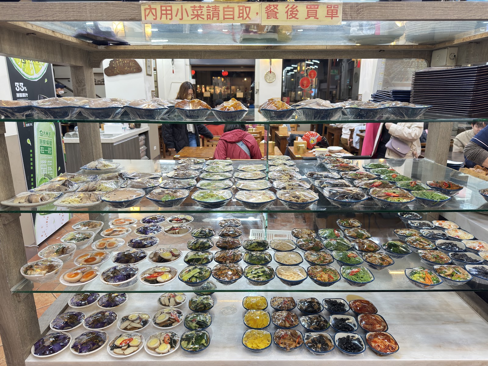 十一街小菜展示架，頂部寫著內用小菜請自取餐後買單，多層玻璃架上排滿各式小菜