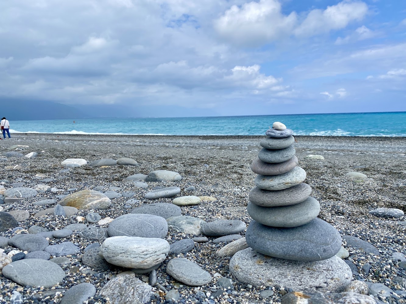 前景有堆疊石頭，背景為藍海與遠方散步旅人的七星潭海灘