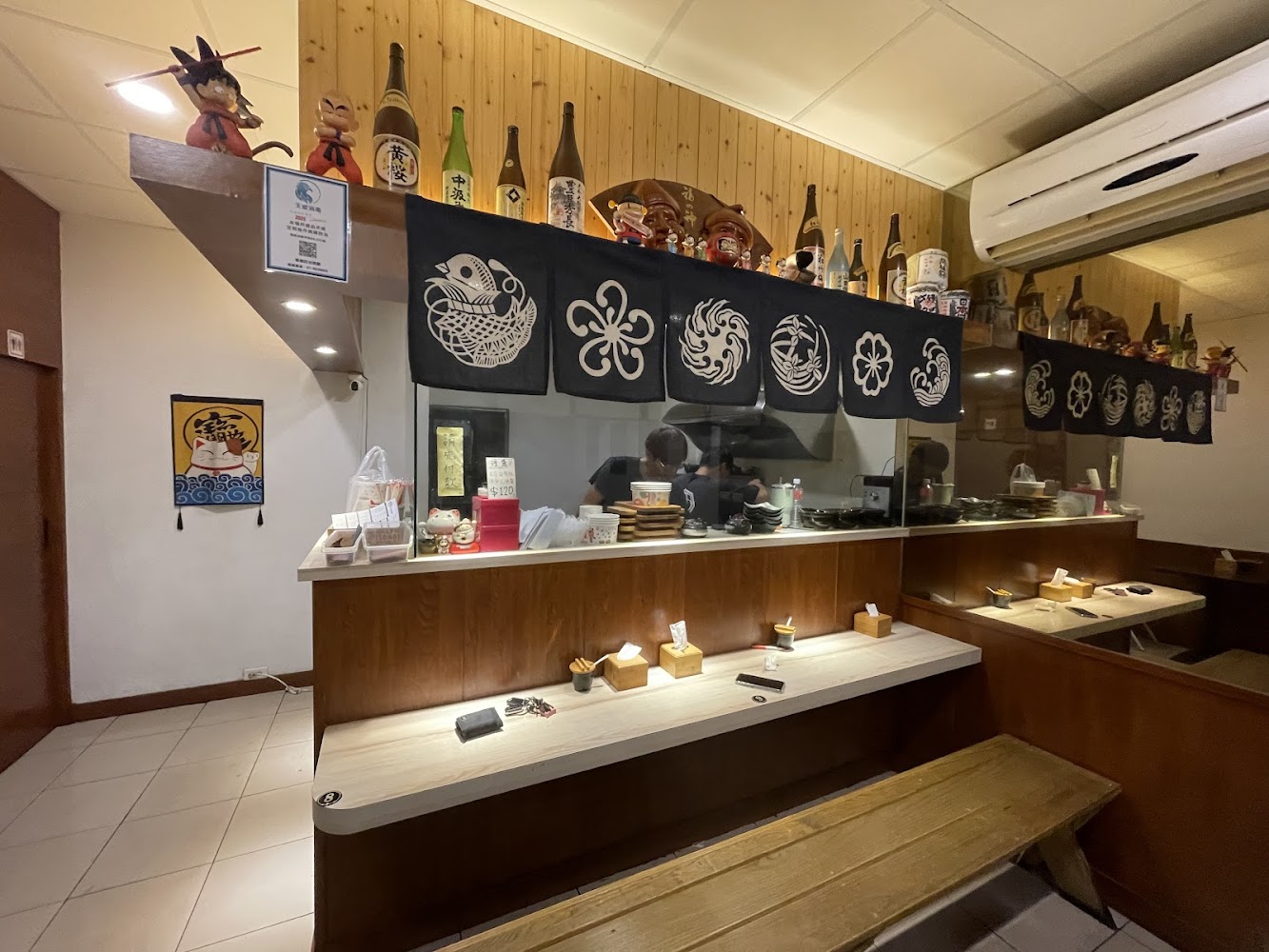 這張圖片主要是日本風格餐廳的吧檯區，有擺放日本清酒瓶、動漫公仔、和式布幔，以及一些用餐座位，整體氛圍溫暖且充滿日式特色。