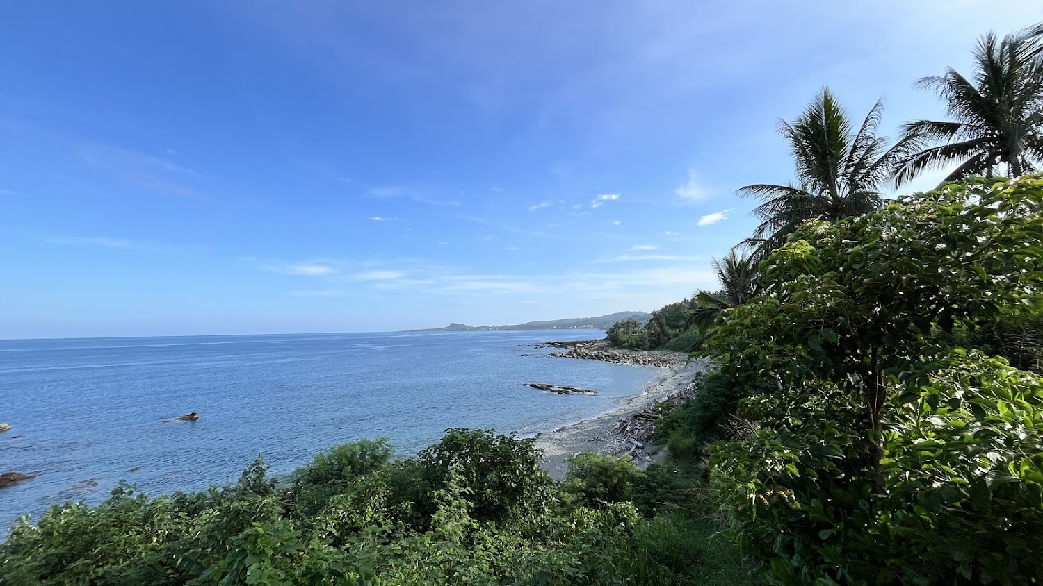 一片湛藍的海洋，沿岸有石頭及小沙灘，周圍綠樹叢生，還有椰子樹
