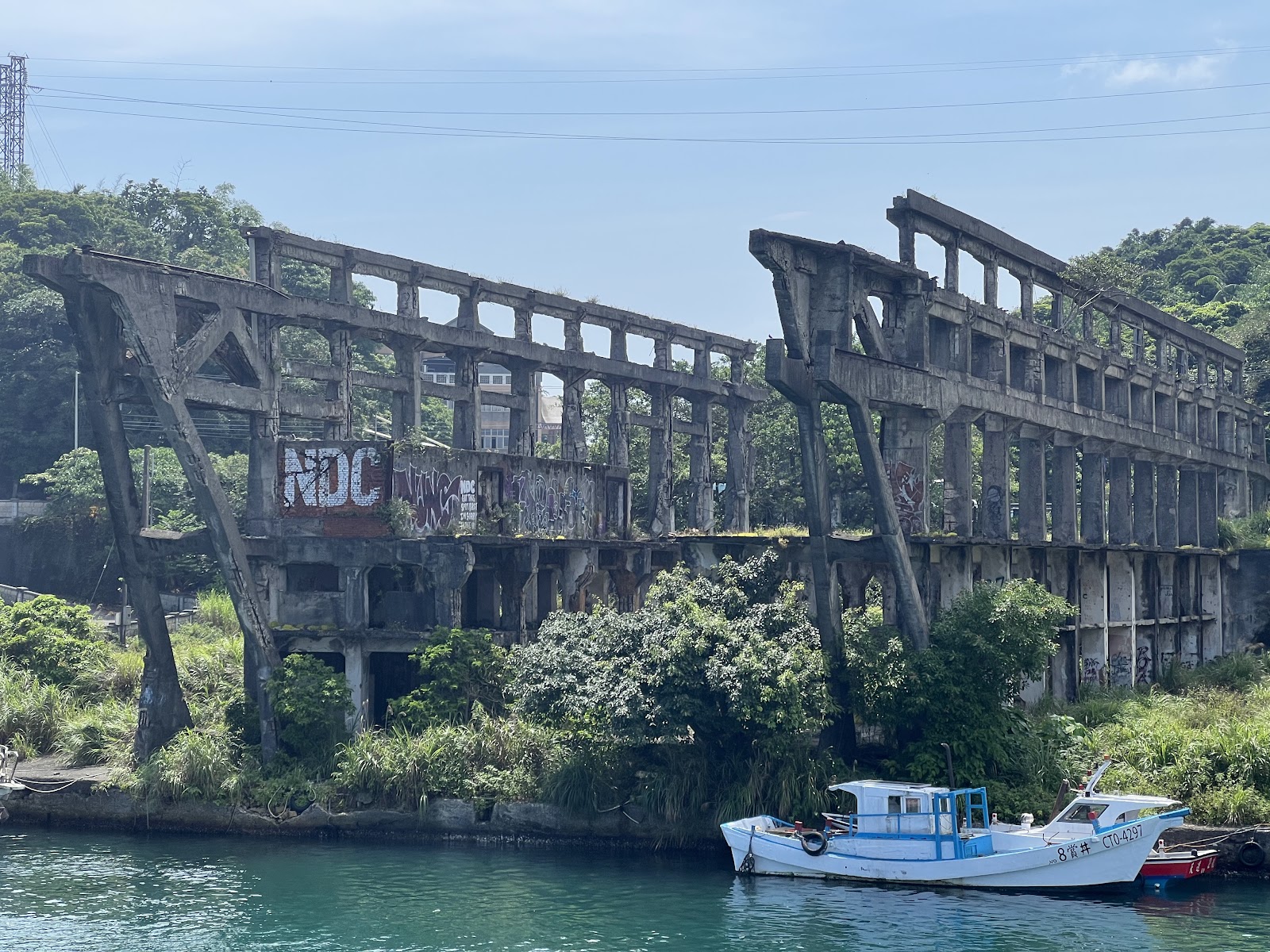 阿根納造船廠廢棄廠房遺構，鏽蝕混凝土骨架覆蓋植披，下方有漁船停靠