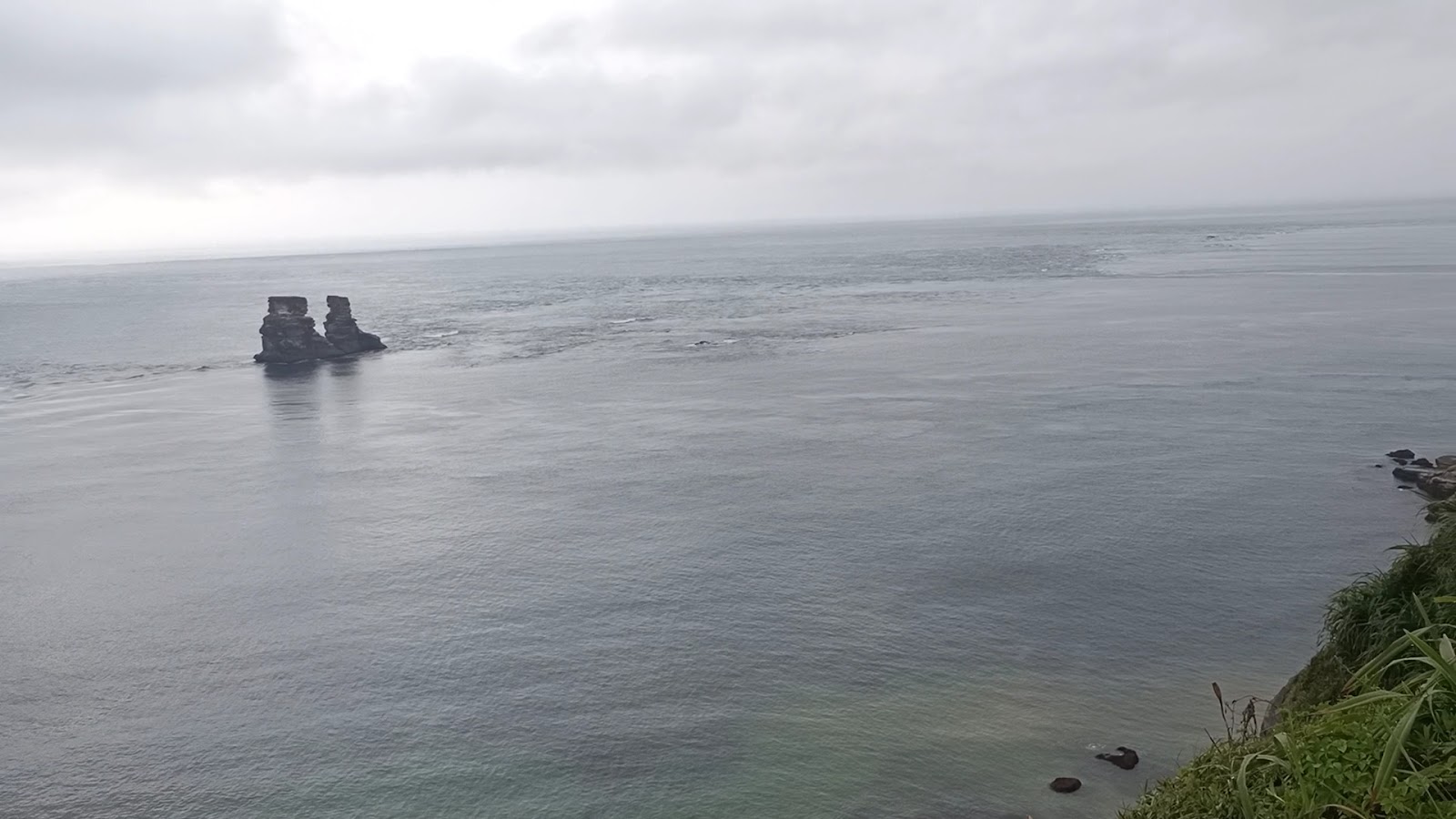 從崖頂俯瞰燭台雙嶼雙礁，渺小矗立灰色海面，崖邊青草，陰天雨霧
